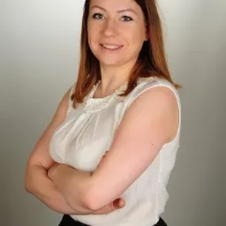 Portret uśmiechniętej kobiety z brązowymi włosami, ubrana w białą bluzkę z ozdobnym dekoltem i czarną spódnicę, z założonymi rękami na neutralnym tle.