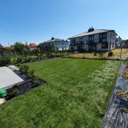 Widok z góry na świeżo rozłożony trawnik z rolki w ogrodzie, z widocznymi fragmentami siatki zabezpieczającej, systemem nawadniania, nowymi nasadzeniami roślin oraz nowoczesnymi domami w tle...