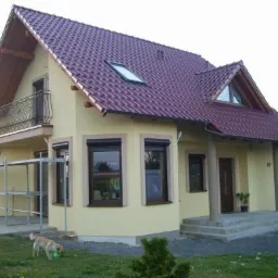 Żółty dom jednorodzinny z czerwoną dachówką widziany z zewnątrz, z balkonem i częściowo rozstawionym rusztowaniem obok, pies bawi się na trawniku.