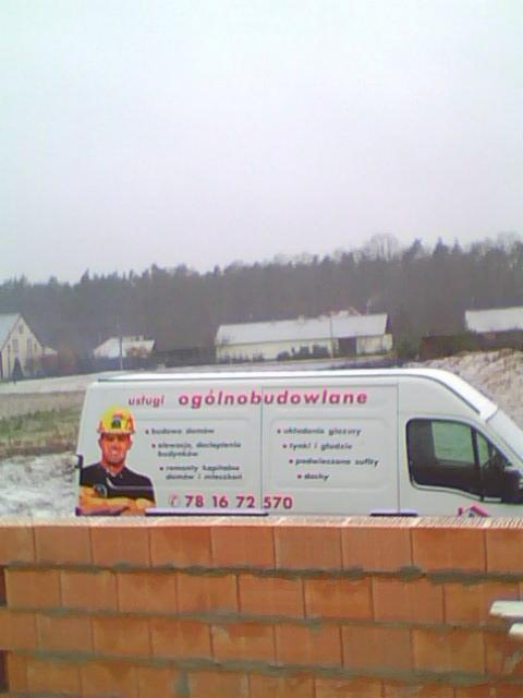 Reklama usług ogólnobudowlanych na samochodzie dostawczym, widoczna zza muru z cegły ceramicznej.