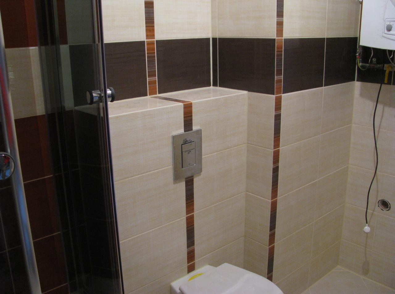 Wykończona łazienka z brązowo-beżowymi płytkami, widoczna kabina prysznicowa z chromowanymi elementami, podwieszana toaleta i bojler elektryczny na ścianie.