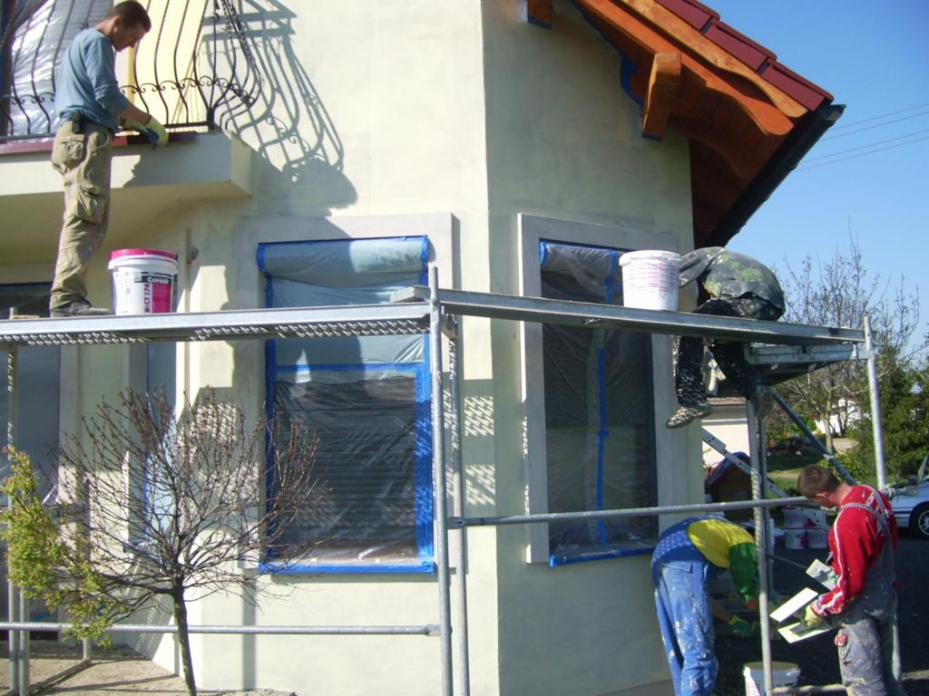 Trzech mężczyzn na rusztowaniu maluje elewację domu, okna zabezpieczone folią i taśmą, widoczne puszki z farbą i narzędzia malarskie.