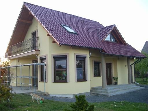 Żółty dom jednorodzinny z czerwoną dachówką widziany z zewnątrz, z balkonem i częściowo rozstawionym rusztowaniem obok, pies bawi się na trawniku.