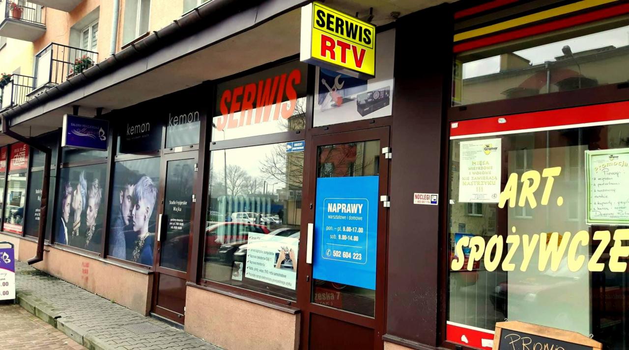 Fasada budynku z kilkoma lokalami usługowymi: Serwis RTV z żółtym szyldem, salon fryzjerski 'Kemon' z grafikami na oknach i sklep spożywczy 'Art. Spożywcze' z promocjami na szybie.