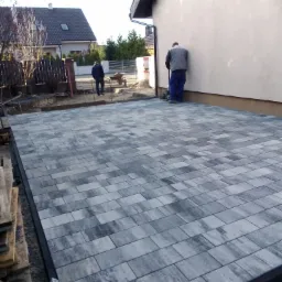 Ułożona szara kostka brukowa na posesji, widoczny pracownik docinający element przy ścianie budynku, drugi pracownik z taczką w tle.