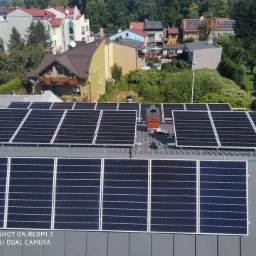 Instalacja paneli na szarym dachu budynku z kominem, widok z g&oacute;ry na tle zabudowy mieszkalnej i zieleni.