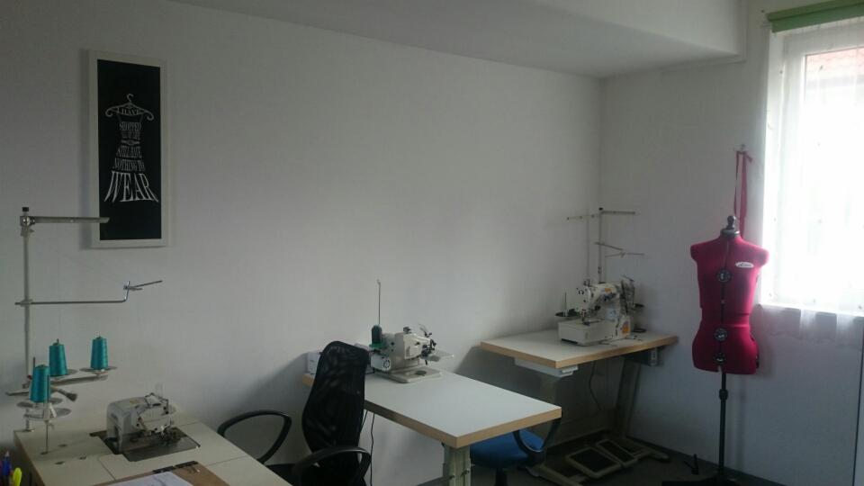 Wnętrze pracowni krawieckiej z maszynami do szycia, manekinem krawieckim w kolorze czerwonym, biurkami, krzesłami i czarną tablicą z napisem 'Nothing to wear'.