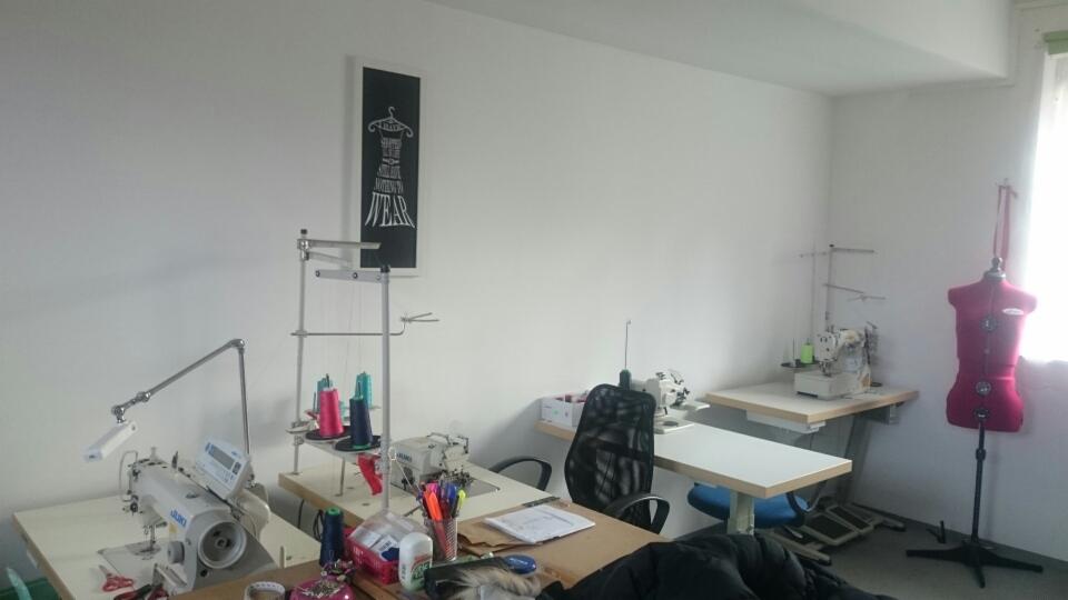 Jasne wnętrze pracowni krawieckiej z maszynami do szycia, manekinem krawieckim w kolorze fuksji, stołami roboczymi, niciami i plakatem z motywem sukienki na ścianie.