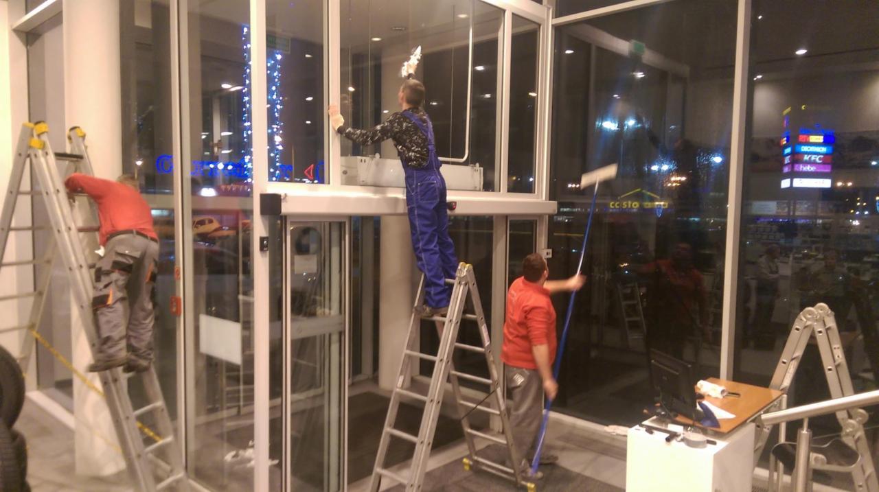 Trzech mężczyzn na drabinach myje duże okna w budynku z widokiem na nocne miasto i szyldy reklamowe.