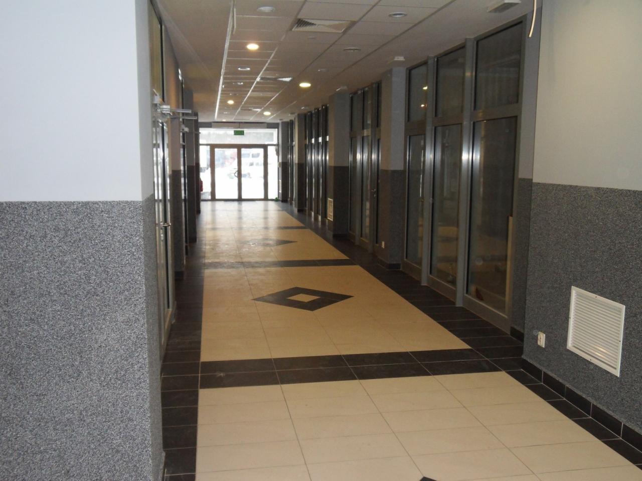 Wnętrze długiego korytarza biurowego z szarymi ścianami wykończonymi imitacją kamienia, jasną podłogą z ciemnymi akcentami i rzędem przeszklonych biur po prawej stronie, widok w kierunku...