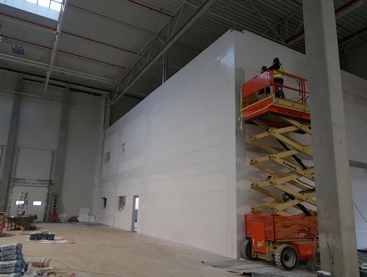 Dwóch pracowników na podnośniku koszowym, malujących białą ścianę wewnątrz hali przemysłowej, widoczne worki z zaprawą i otwarte drzwi w tle.