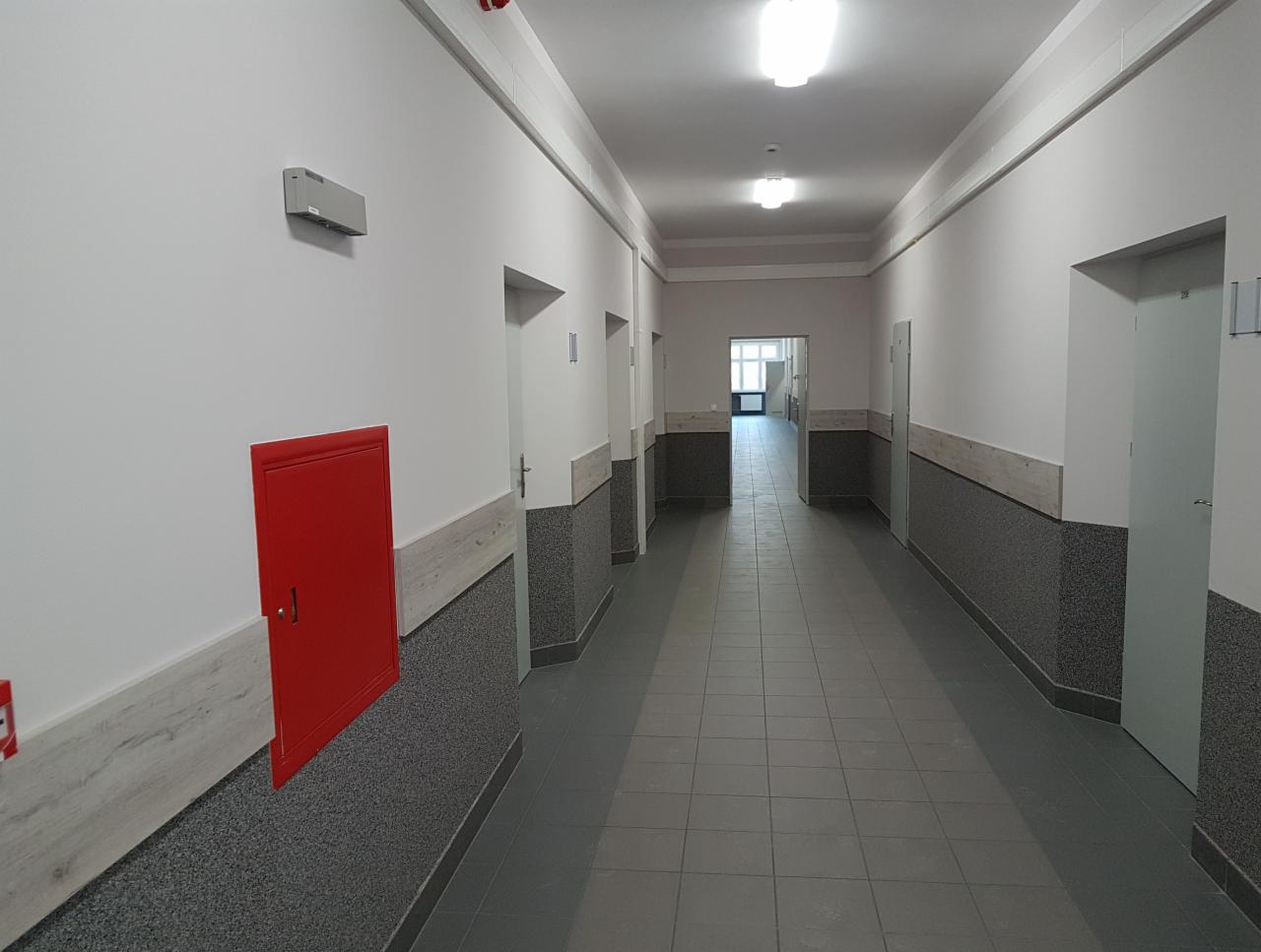 Długi korytarz biurowy z szarą podłogą, biało-szarymi ścianami i jaskrawoczerwoną szafką hydrantową po lewej stronie.
