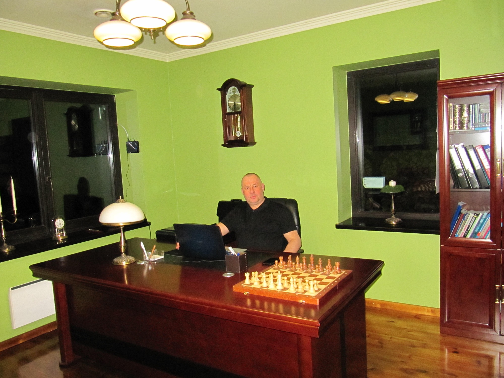 Mężczyzna siedzi przy biurku z laptopem i szachami w zielonym gabinecie z biblioteczką.
