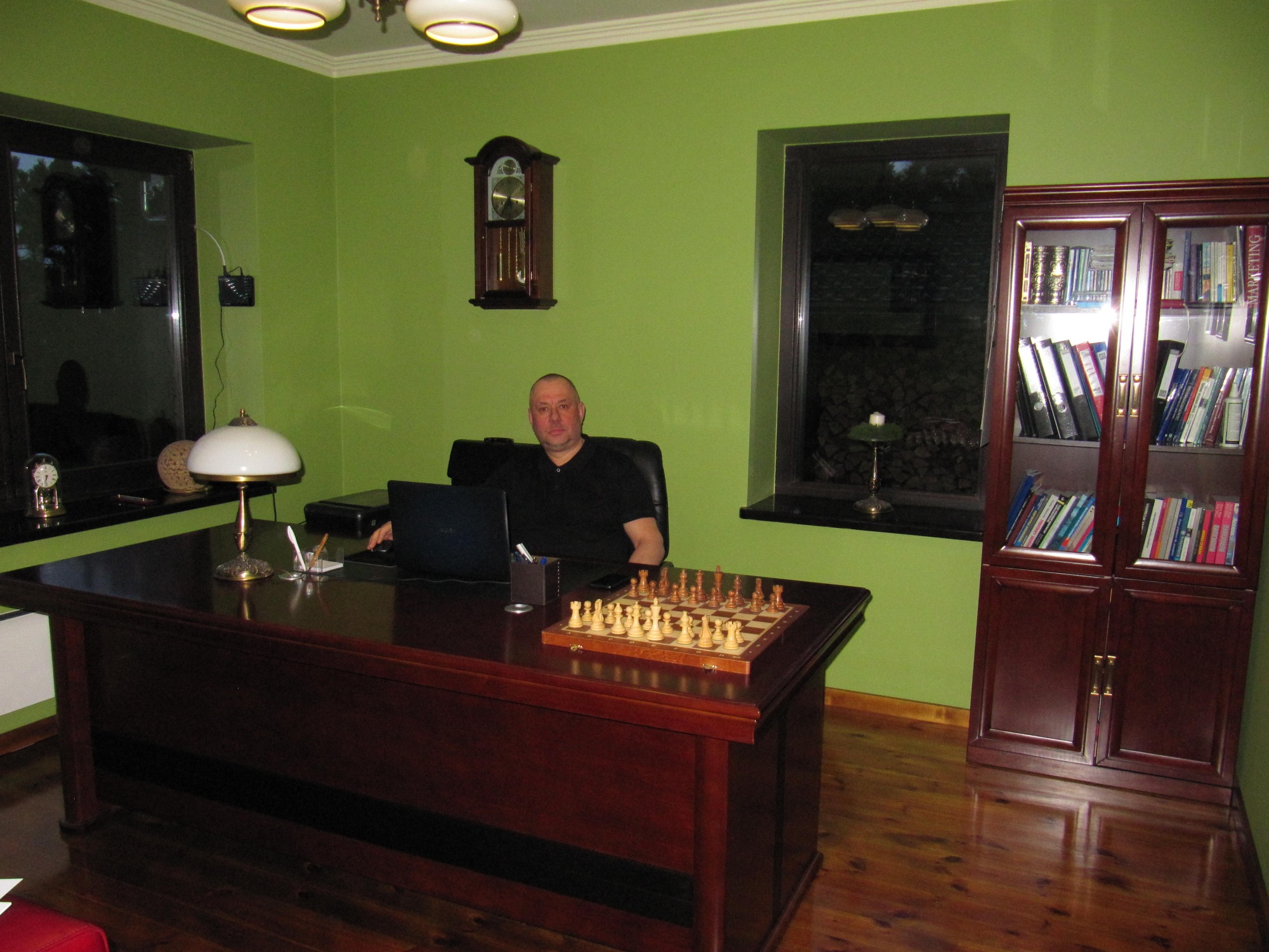 Mężczyzna w czarnej koszulce siedzi za dużym, mahoniowym biurkiem z laptopem i szachami w gabinecie o zielonych ścianach.