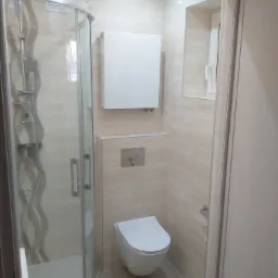 Jasna łazienka po remoncie z kabiną prysznicową z wzorem fali na szklanej ściance, podwieszaną toaletą i grzejnikiem na ścianie wyłożonej beżowymi płytkami imitującymi drewno.