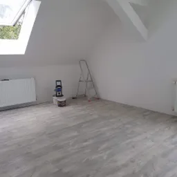 Poddasze po remoncie z nową, jasną podłogą imitującą drewno, świeżo pomalowanymi na biało ścianami, grzejnikiem, oknem dachowym i rozstawioną drabiną malarską w rogu.