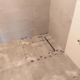 Układanie płytek podłogowych w łazience, widoczny odpływ liniowy i niebieskie krzyżyki dystansowe, jasne płytki imitujące beton.