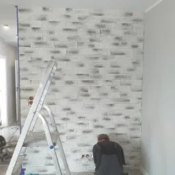 Pracownik w kombinezonie montuje dekoracyjną okładzinę ścienną imitującą kamień na ścianie w szarym pomieszczeniu, widoczna drabina, narzędzia i fragmenty okładziny na podłodze.