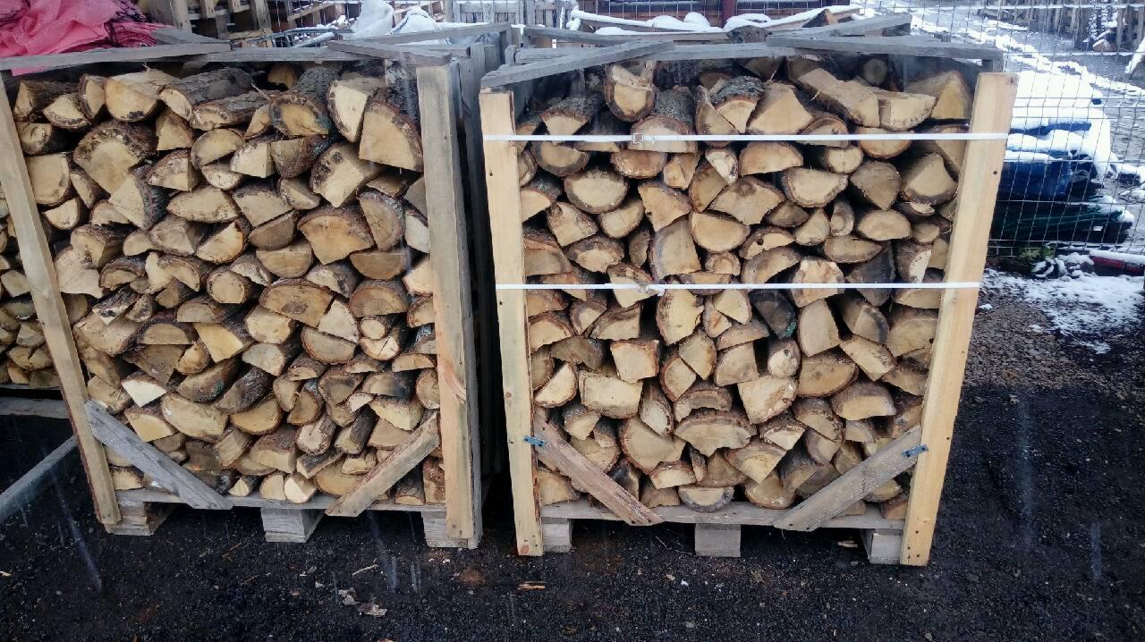 Dwa palety z drewnem kominkowym, zabezpieczone białym sznurkiem, ustawione na zewnątrz, na ziemi z resztkami śniegu.