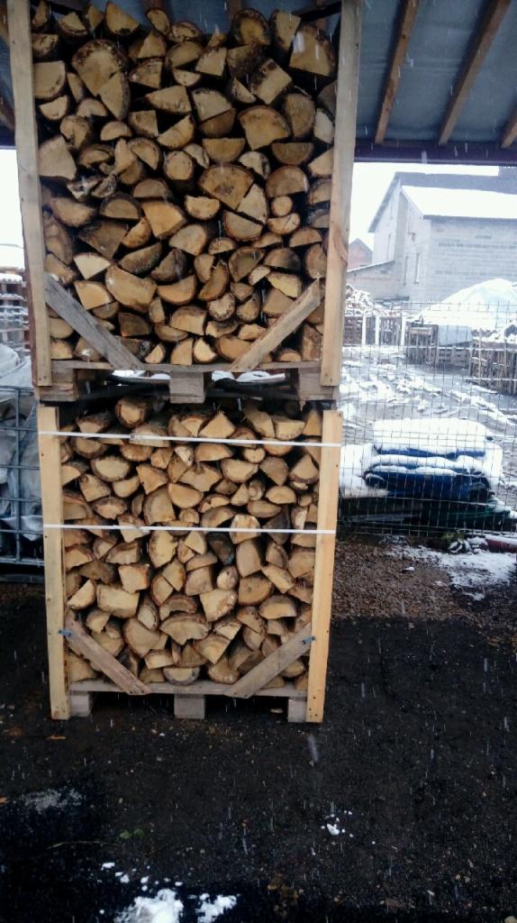 Dwa drewniane kontenery ułożone jeden na drugim, wypełnione pociętym drewnem kominkowym, zabezpieczone białym sznurkiem, na tle zimowego krajobrazu z padającym śniegiem.