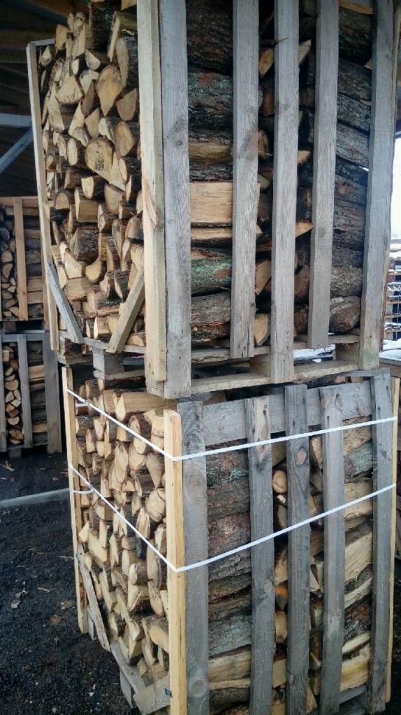 Dwa palety drewna kominkowego ułożone jeden na drugim, zabezpieczone białą liną, widoczne pod zadaszeniem.