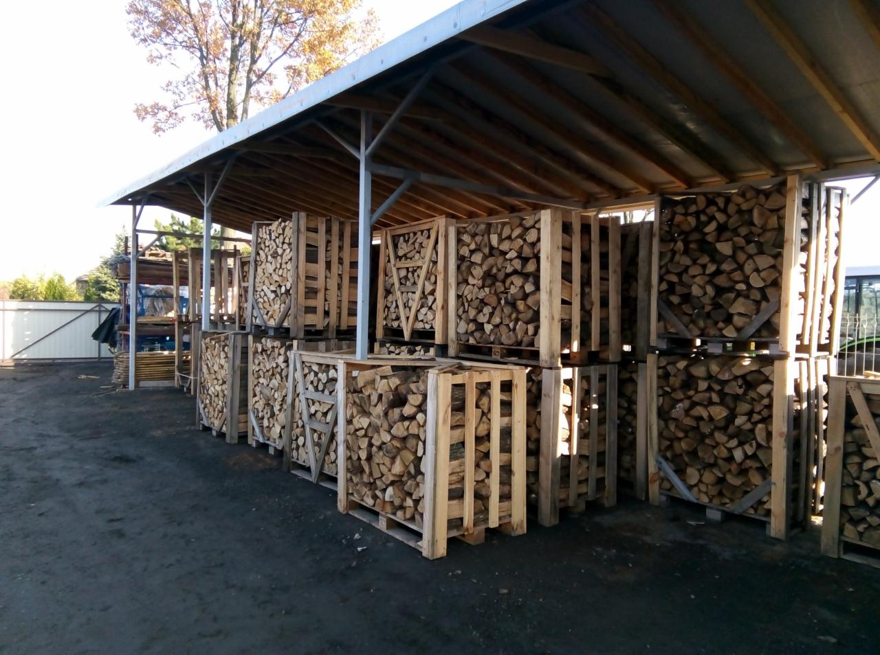 Ułożone palety z pociętym drewnem kominkowym pod zadaszeniem, widoczne drzewa w tle.