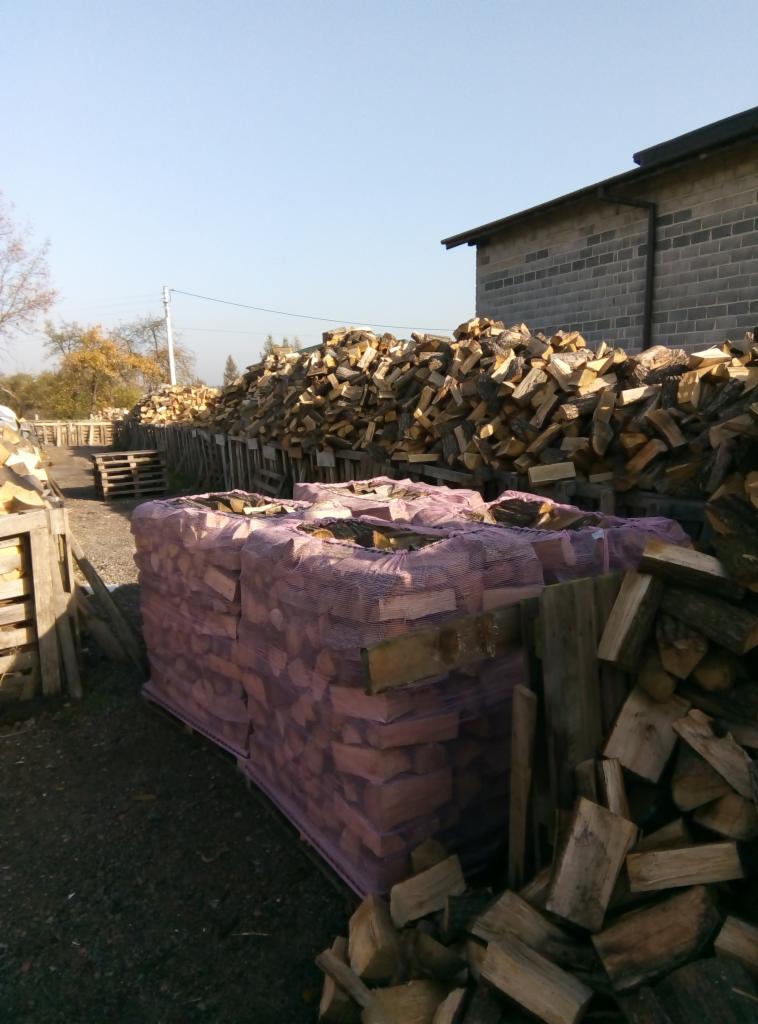 Składowane drewno kominkowe: pocięte i ułożone w paletach z siatką, z widocznymi stosami drewna w tle i budynkiem z szarej cegły.