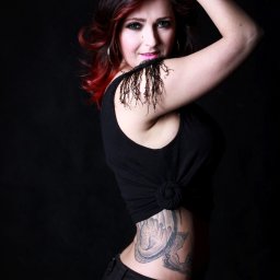 Jakubowska FOTO - Kobieta z czerwoną czupryną, w czarnym topie z frędzlami i tatuażem smoka na boku, pozująca na czarnym tle.