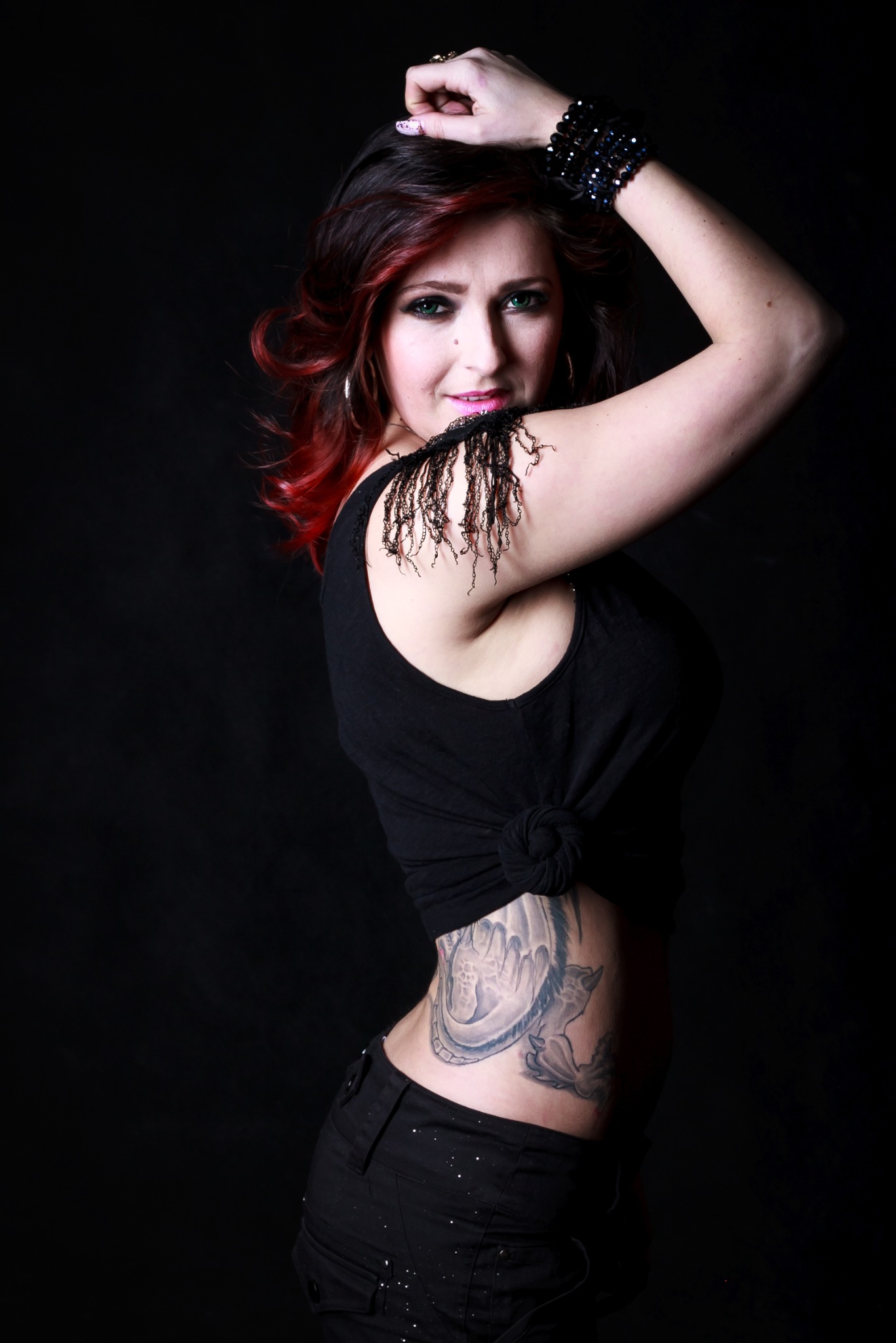 Kobieta z czerwoną czupryną, w czarnym topie z frędzlami i tatuażem smoka na boku, pozująca na czarnym tle.