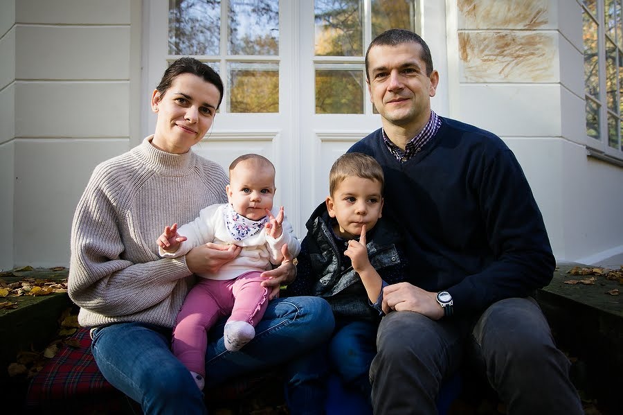 Rodzinne zdjęcie: mama trzyma niemowlę w różowych spodniach, tata z synkiem siedzą na tle białych drzwi i jesiennego ogrodu.