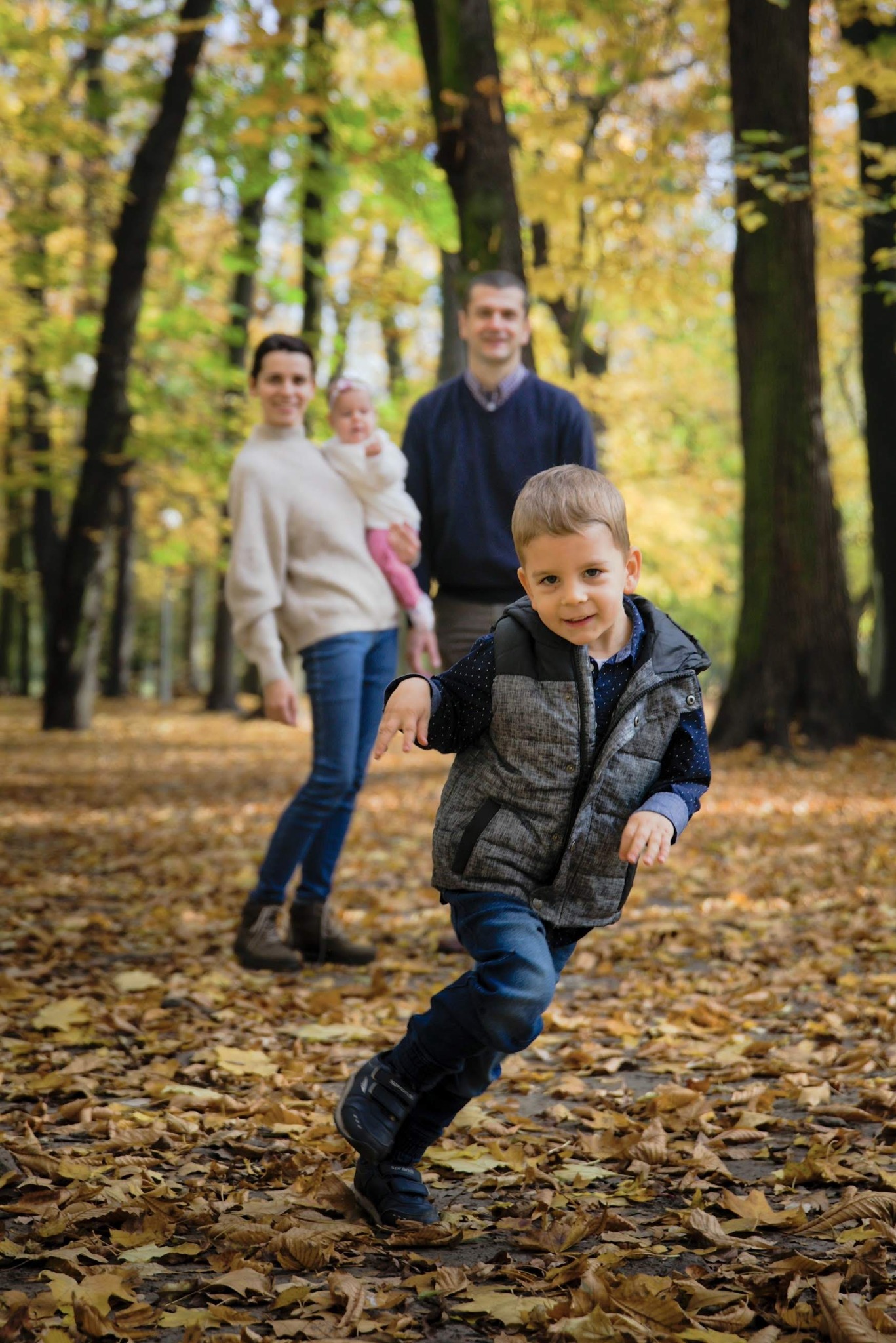 Chłopiec biegnie pośród jesiennych liści, w tle rodzina z niemowlęciem w parku.