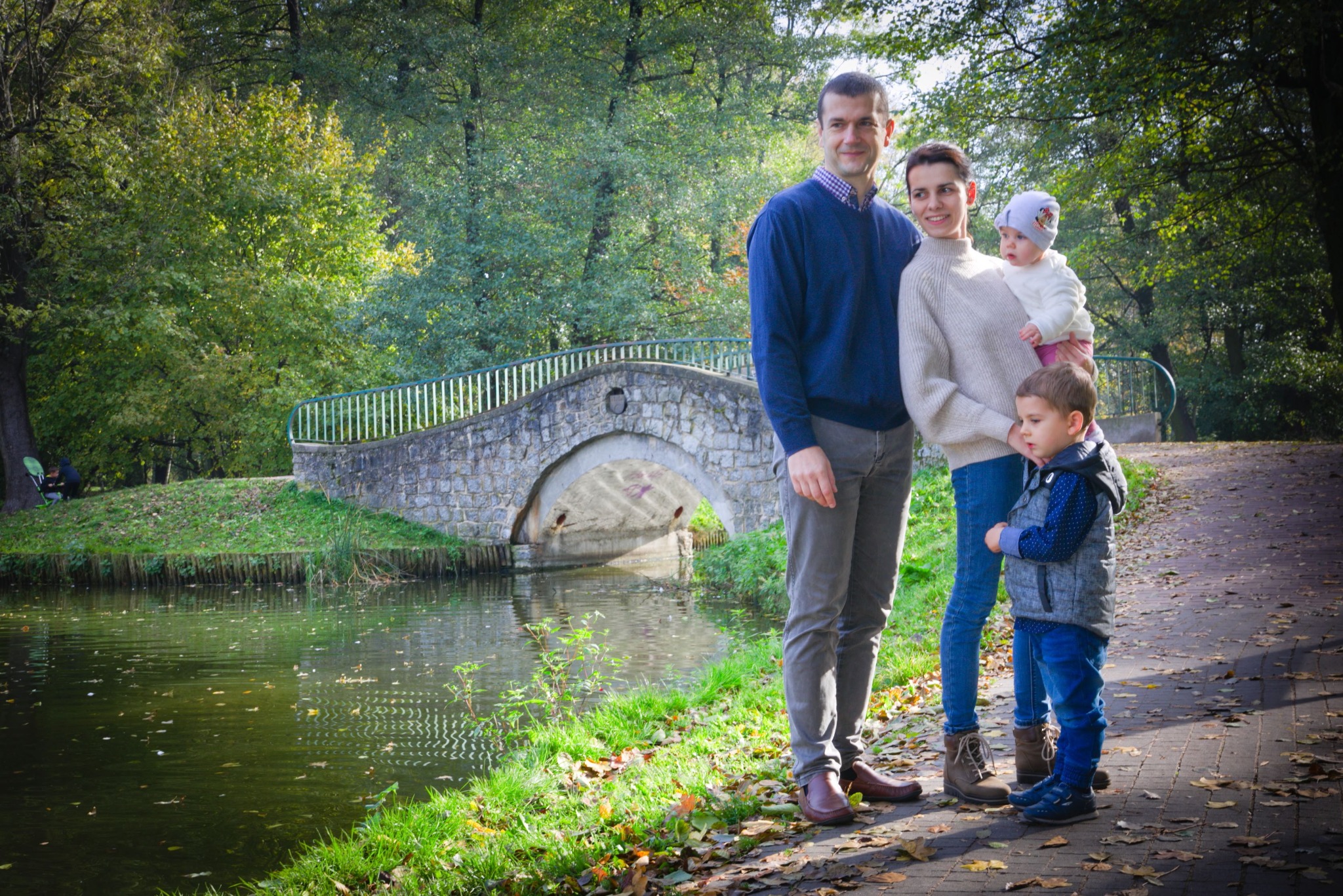 Rodzina z dwójką dzieci pozuje na tle kamiennego mostku w parku, jesienne liście na ścieżce.