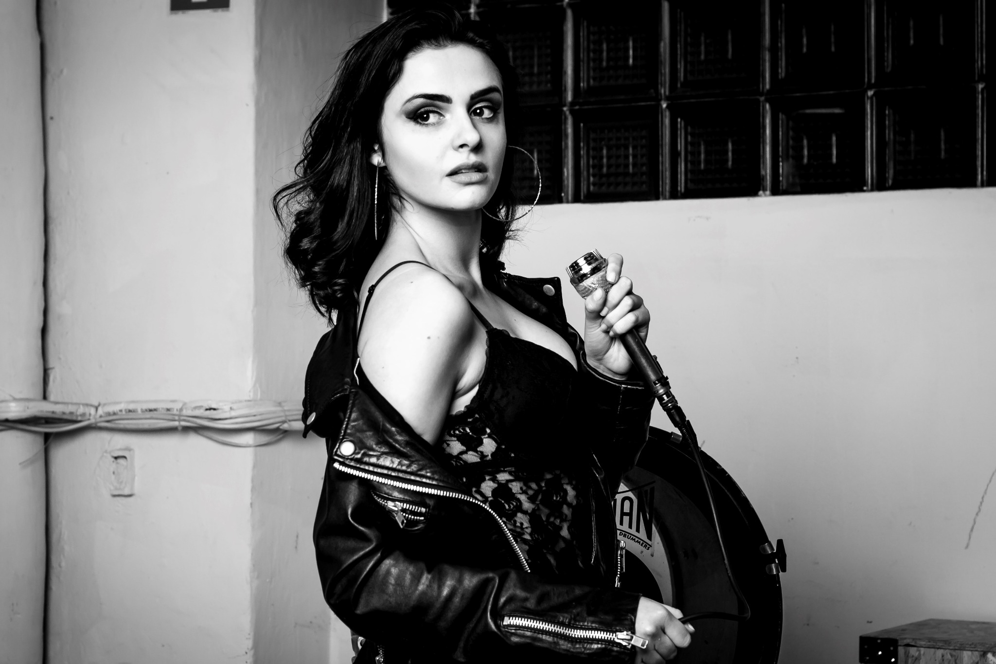 Czarno-białe zdjęcie kobiety z mikrofonem, ubrana w koronkową bluzkę i skórzaną kurtkę, z perkusją w tle