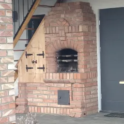 Murowany grill z cegły z elementami drewnianymi, wbudowany pod schodami zewnętrznymi, z widoczną kratką i popielnikiem, obok szara ściana z drzwiami.