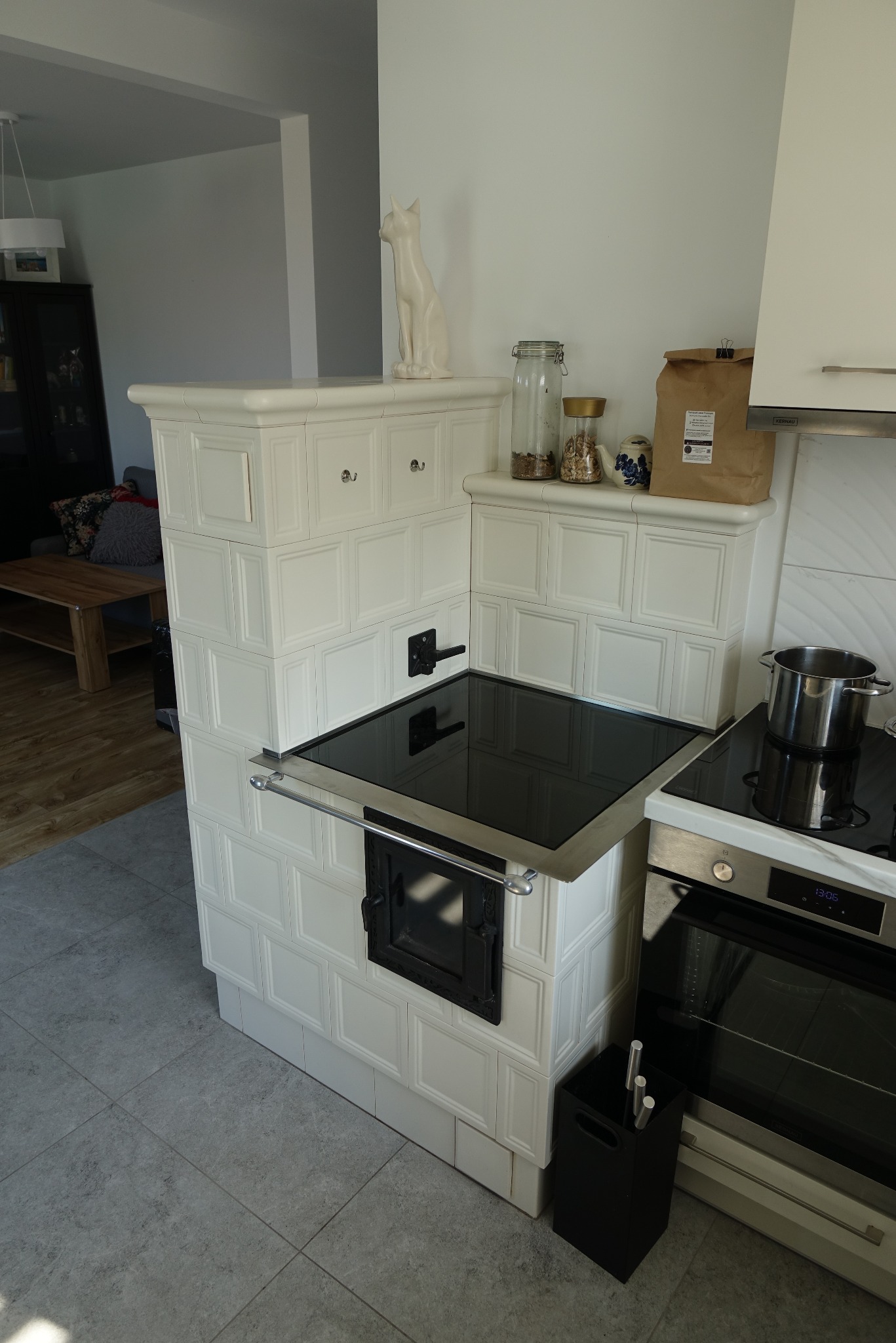 Biały piec kaflowy z płytą grzewczą w nowoczesnej kuchni. Na górze dekoracyjna figurka kota i słoiki. Widoczny fragment piekarnika.