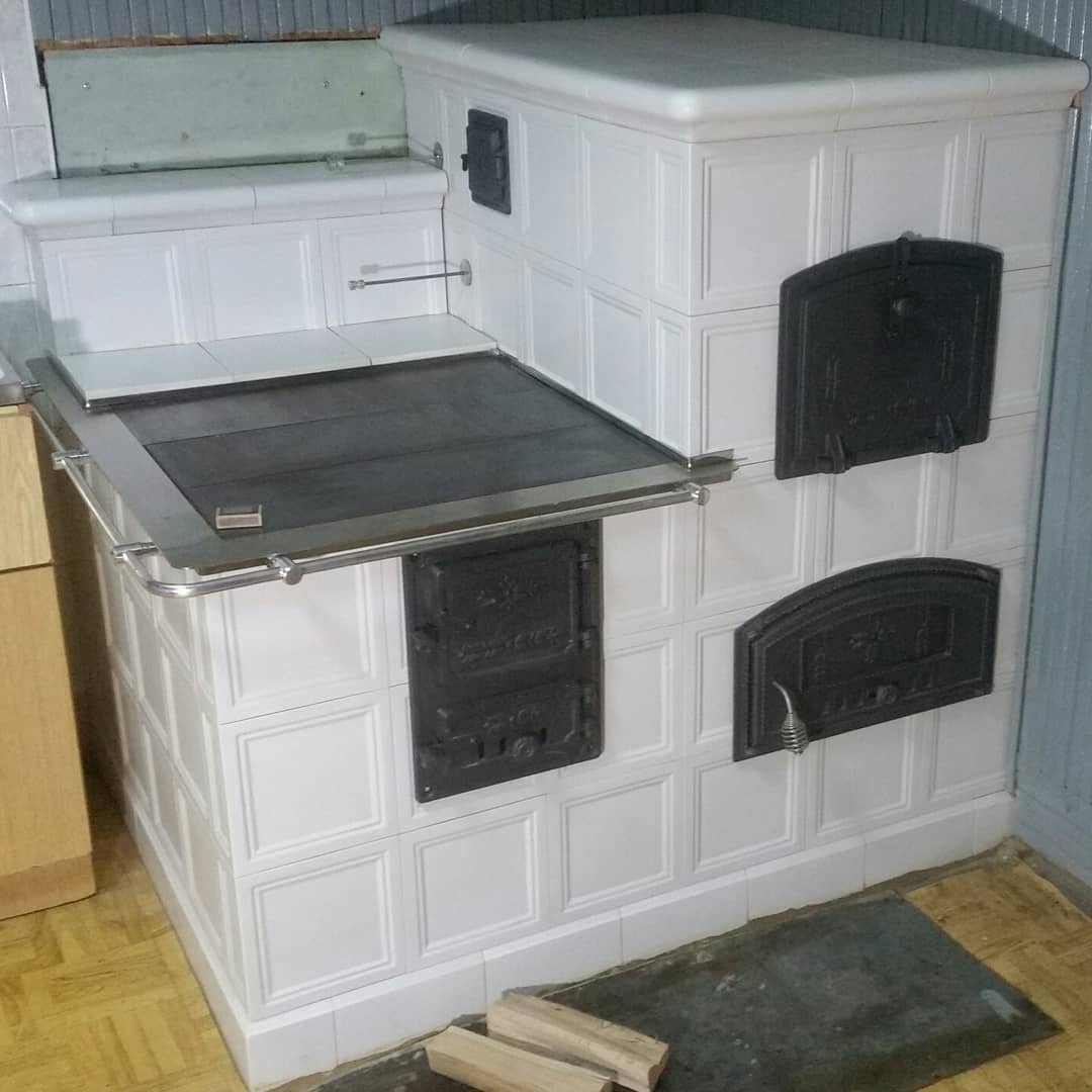 Biały piec kaflowy z żeliwnymi drzwiczkami i blatem kuchennym, widoczne dwa kawałki drewna opałowego na podłodze, stylizacja retro.