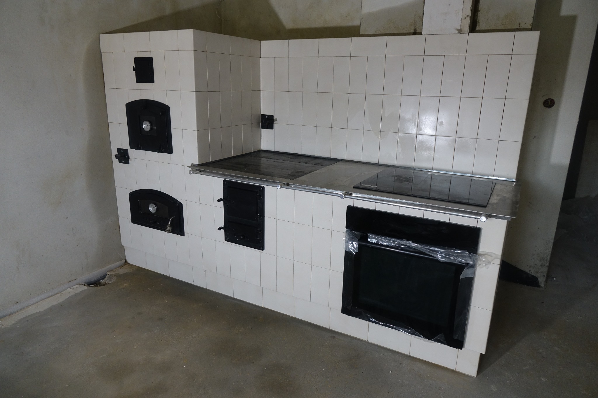 Biała, kafelkowana kuchnia kaflowa z czarnymi drzwiczkami pieca i płytą grzewczą, widoczna folia ochronna na piekarniku.