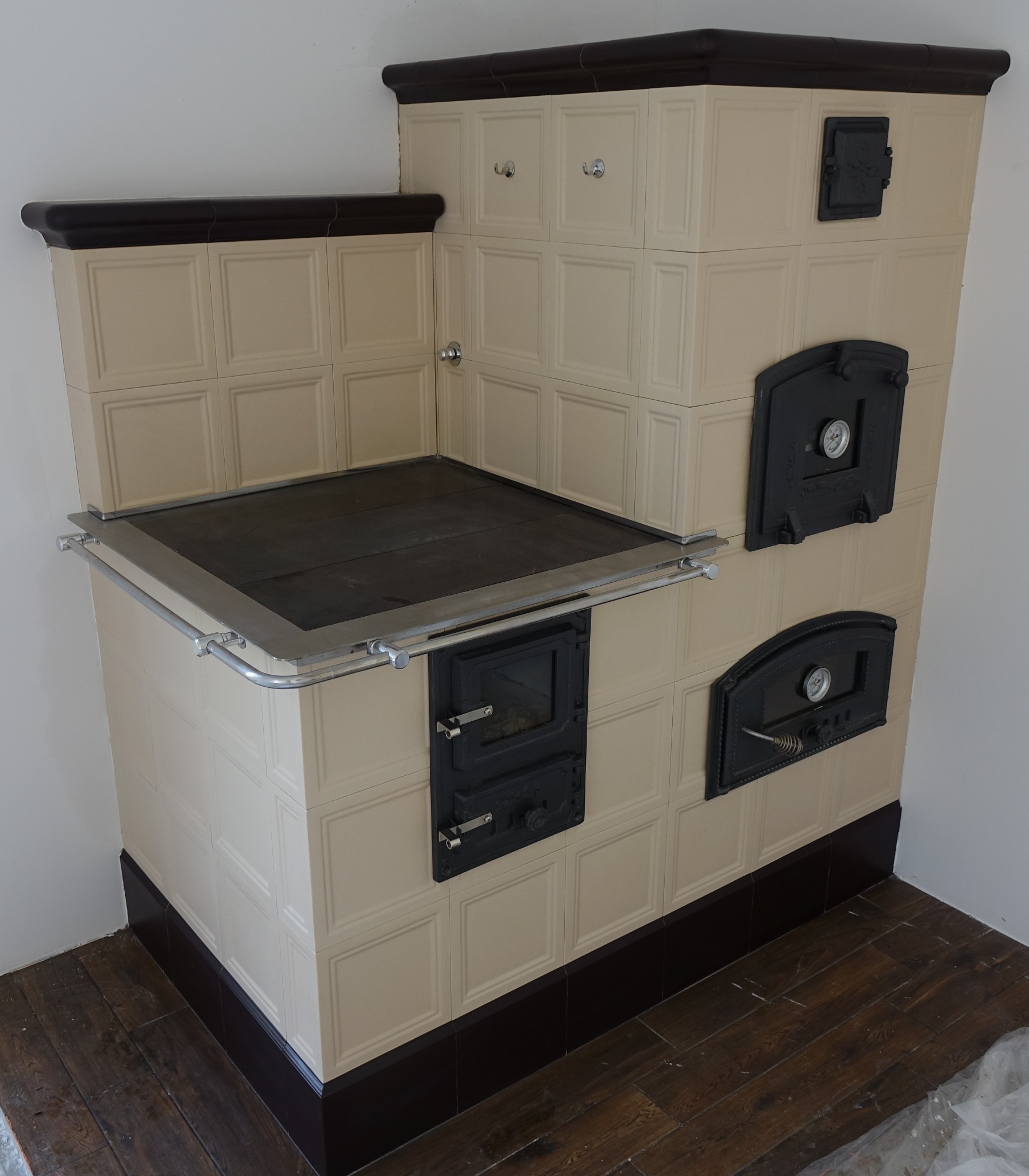 Tylna, boczna perspektywa kaflowego pieca kuchennego z czarnymi drzwiczkami i blatem, wykończonego kremowymi kaflami z ciemnobrązowymi akcentami.