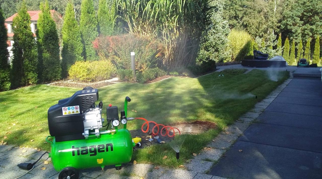 Zielony kompresor Hagen podłączony do instalacji nawadniającej trawnik z dyszami rozpylającymi wodę, widoczne drzewa iglaste w tle.