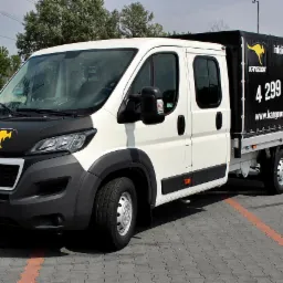 Biało-czarny samoch&oacute;d dostawczy z logo kangura na masce i plandeką z reklamą wypożyczalni Kangoor, zaparkowany na szarym bruku.