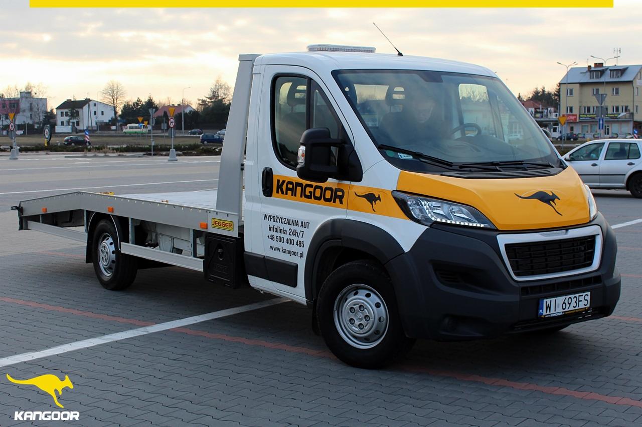 Laweta z platformą załadunkową marki Kangooor na parkingu, z widocznym numerem telefonu do wypożyczalni i logiem kangura.