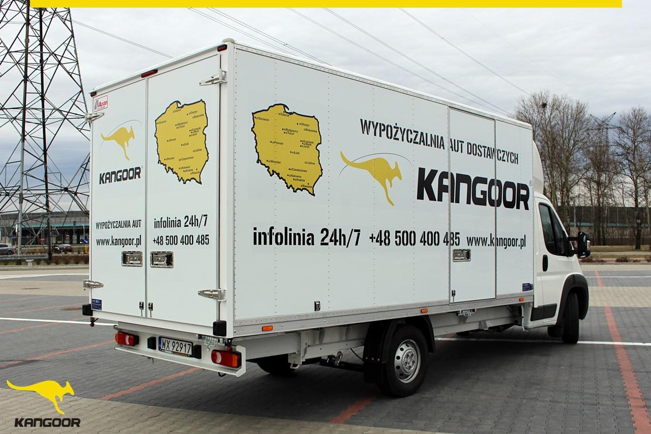 Biała furgonetka z grafiką kangura i mapą Polski, zaparkowana na parkingu, w tle słupy wysokiego napięcia.