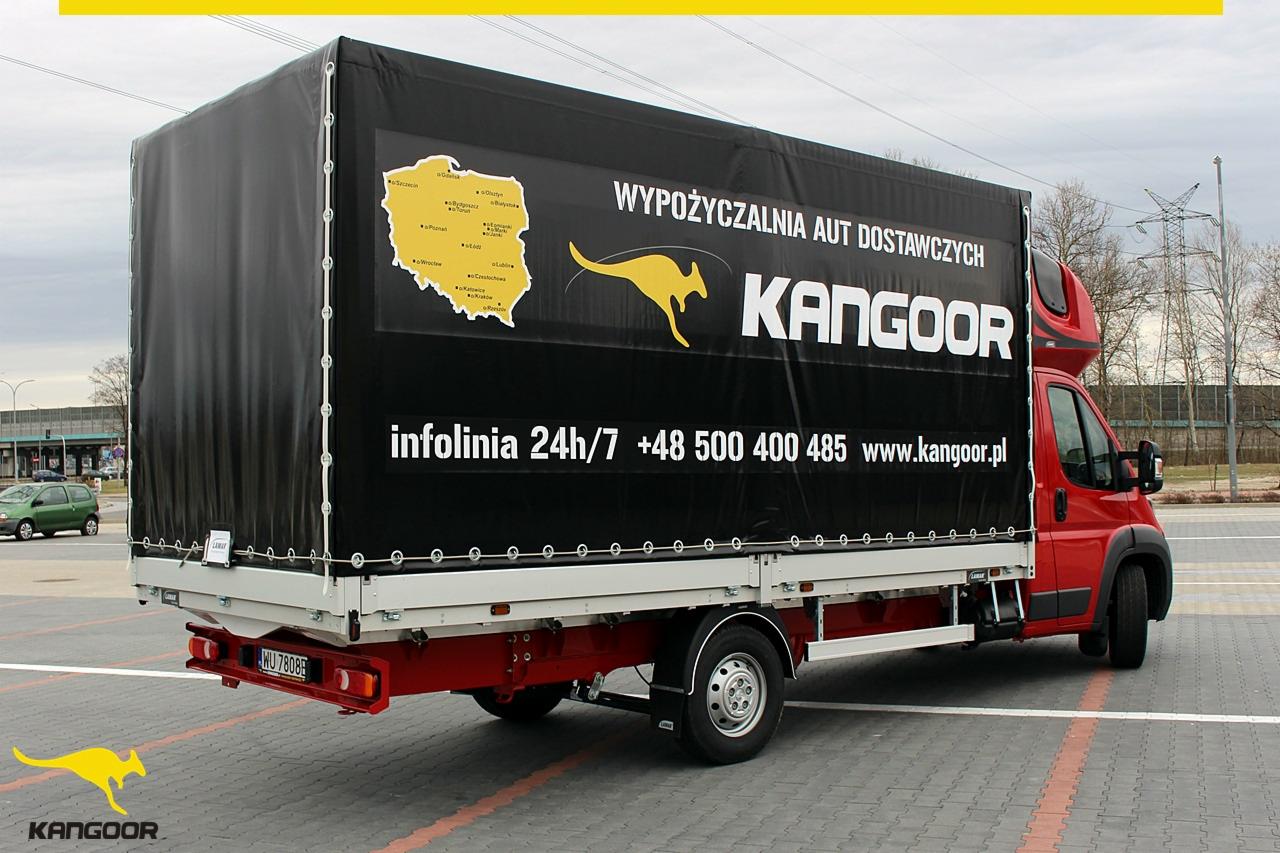 Czerwony samochód dostawczy z czarną plandeką marki KangooR z mapą Polski i numerem infolinii zaparkowany na parkingu.