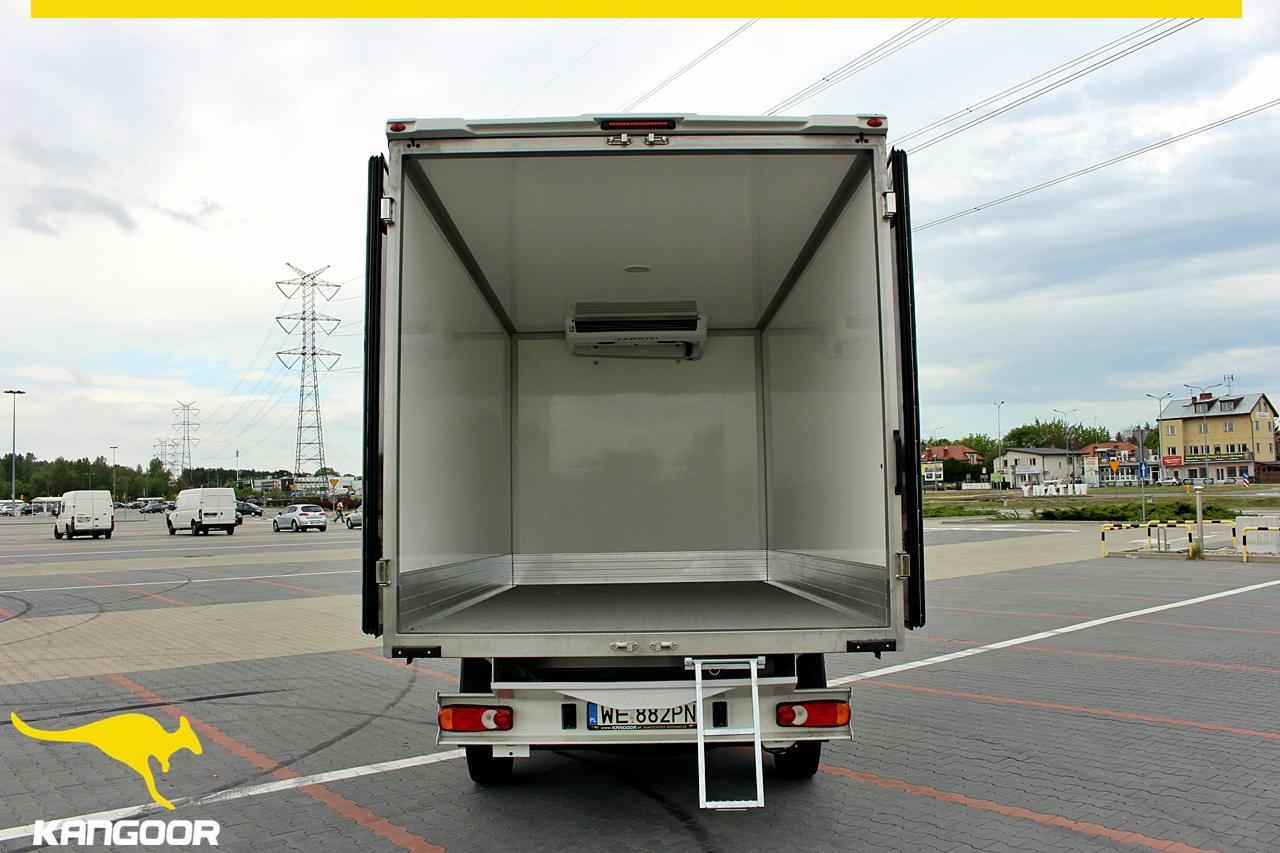 Otwarta pusta przestrzeń ładunkowa chłodni samochodowej z widocznym agregatem chłodniczym, schodkami wejściowymi i numerem rejestracyjnym pojazdu na tle parkingu z liniami energetycznymi w oddali.