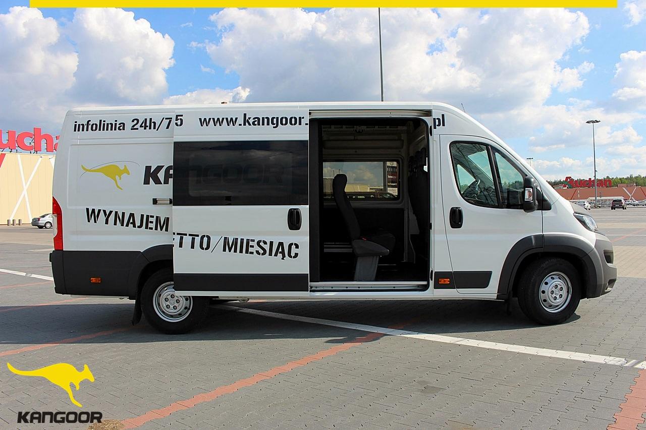 Biały bus z otwartymi drzwiami bocznymi, logo firmy z kangurem i napisem 'Wynajem' na parkingu przed sklepem wielkopowierzchniowym.