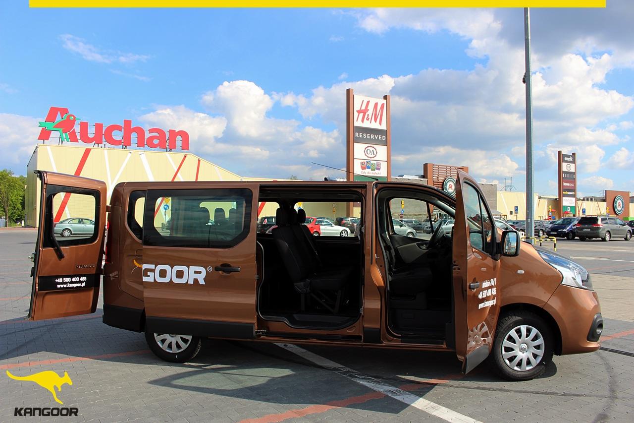 Brązowy van z otwartymi drzwiami na parkingu przed centrum handlowym z widocznymi szyldami Auchan i H&M.