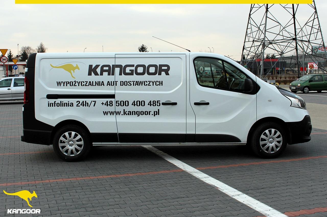 Biały samochód dostawczy z logo firmy i numerem telefonu na parkingu z kostki brukowej, w tle słup energetyczny.