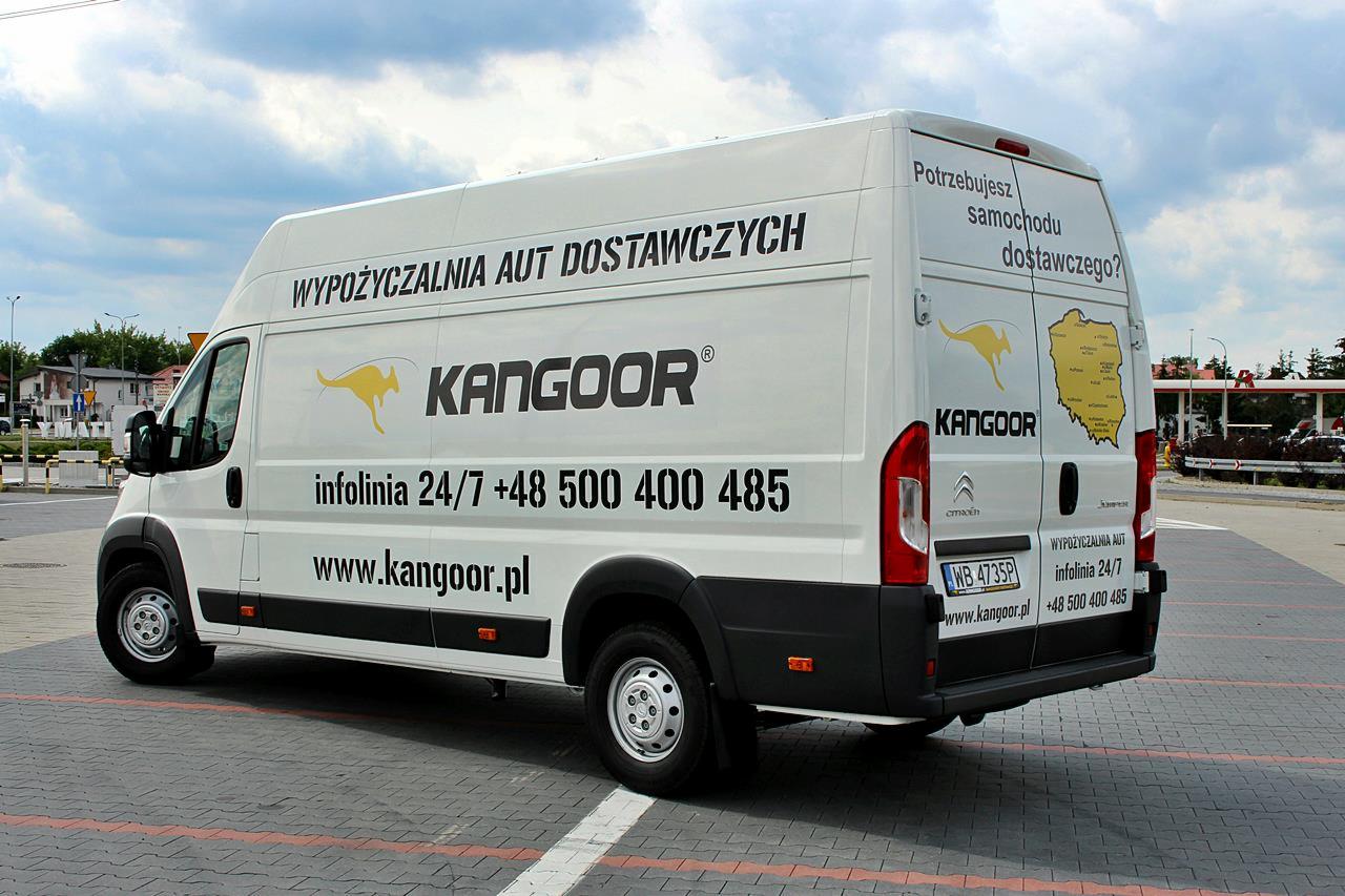Biały samochód dostawczy z logo KangooR i numerem infolinii, zaparkowany na kostce brukowej z widocznym zarysem mapy Polski na tylnych drzwiach.