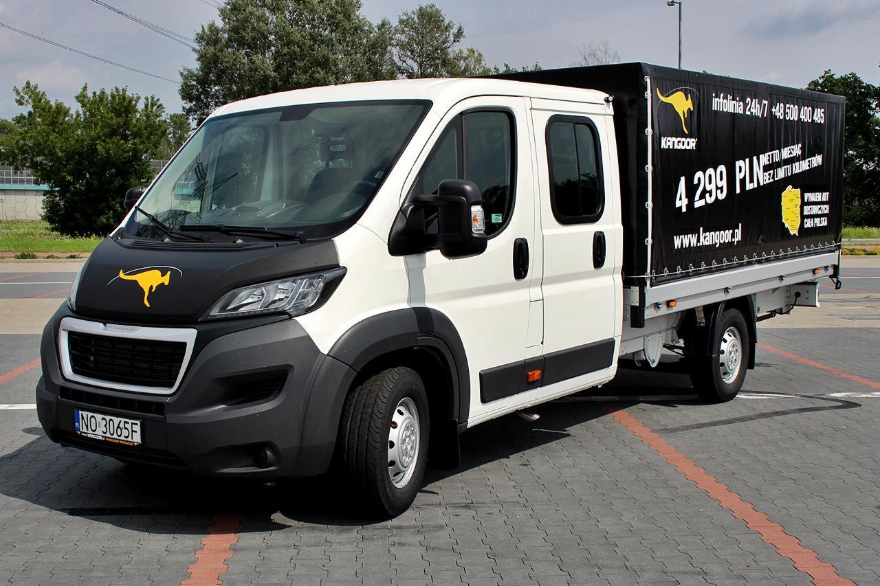 Biało-czarny samochód dostawczy z logo kangura na masce i plandeką z reklamą wypożyczalni Kangoor, zaparkowany na szarym bruku.