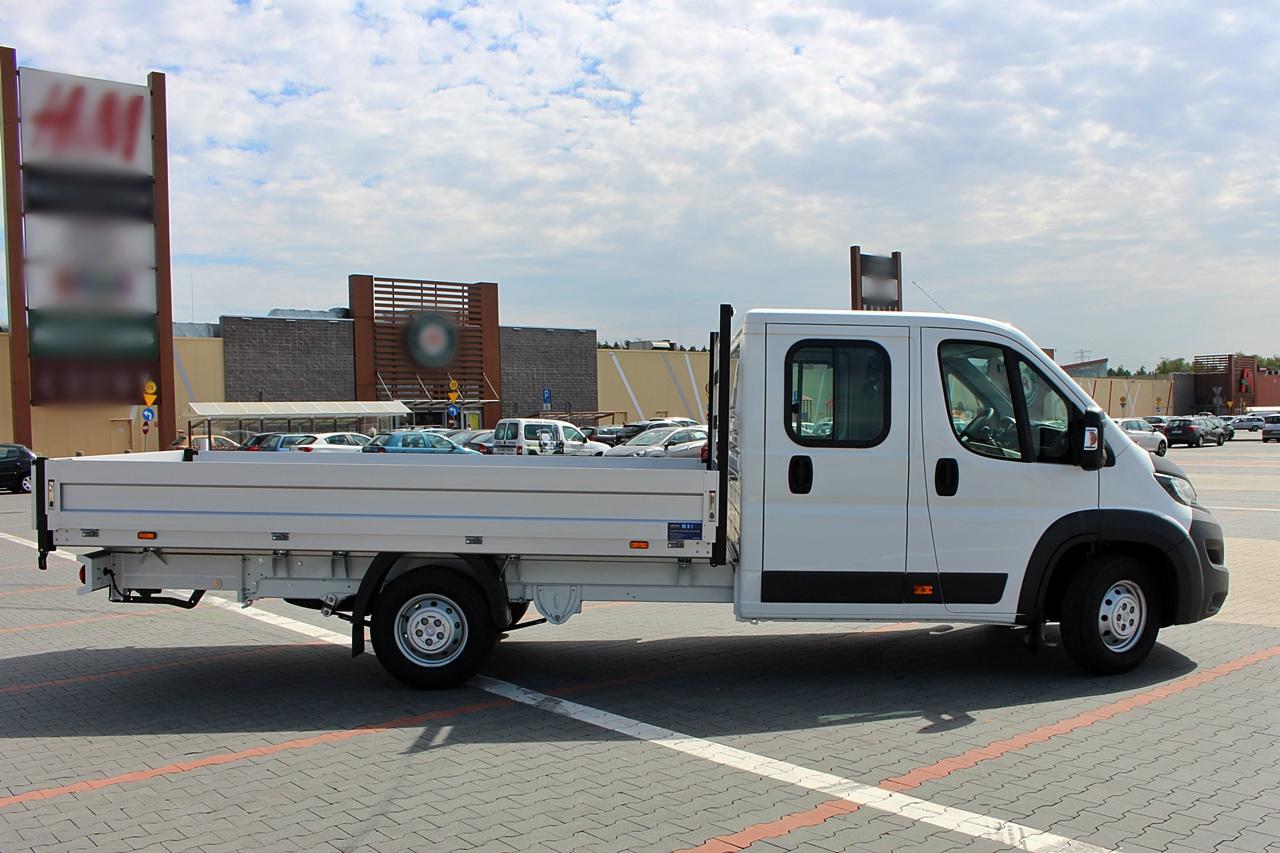 Biały samochód dostawczy z otwartą skrzynią ładunkową na parkingu przed centrum handlowym, widok z boku.
